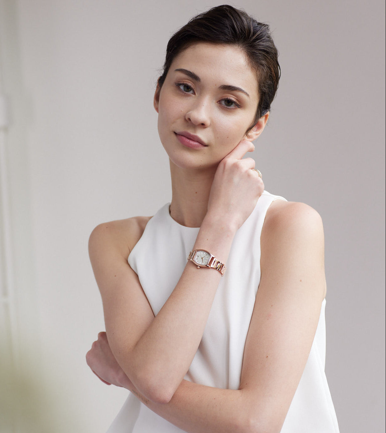 A woman with her hair pulled back wears a white sleeveless top and a rose gold watch with a white dial on her wrist, posing against a soft, light gray background.