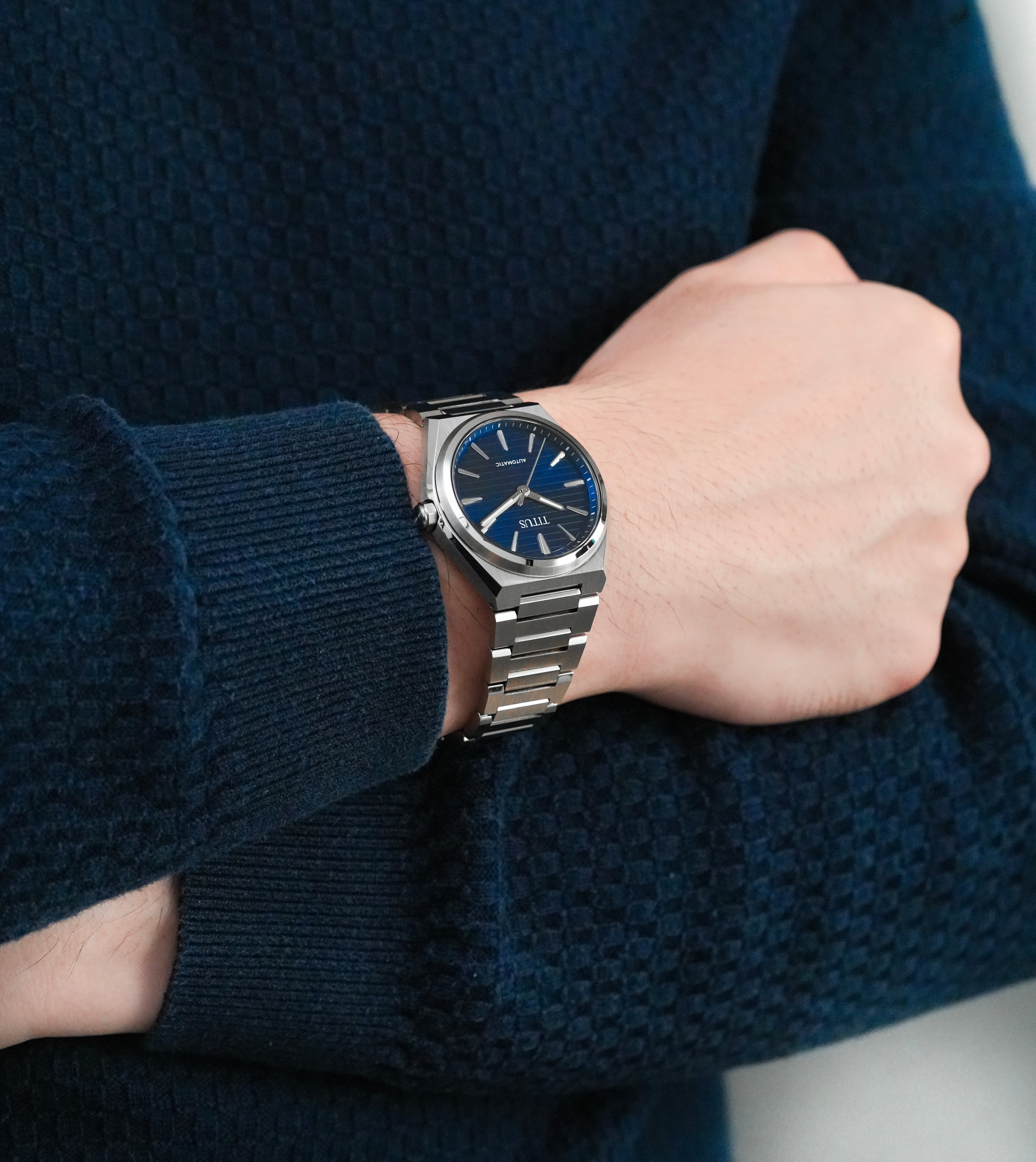 A close-up of a person's wrist wearing a silver-toned metal bracelet watch with a deep blue dial and blue hour markers. The person is wearing a textured navy blue knit sweater.